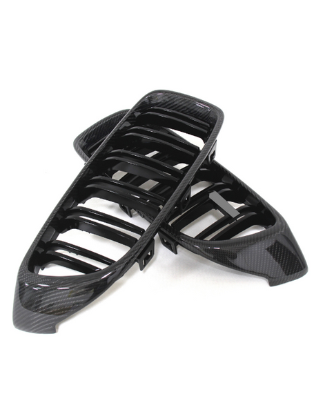 Black car grille covers made of glossy carbon fiber, overlapping each other. They rest on a plain white background, designed for vehicle front-end styling.