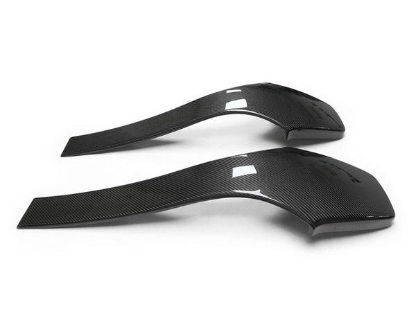 Two curved, carbon fiber car mudguards rest on a plain white surface, displaying a sleek, aerodynamic design with smooth texture and slight reflection.