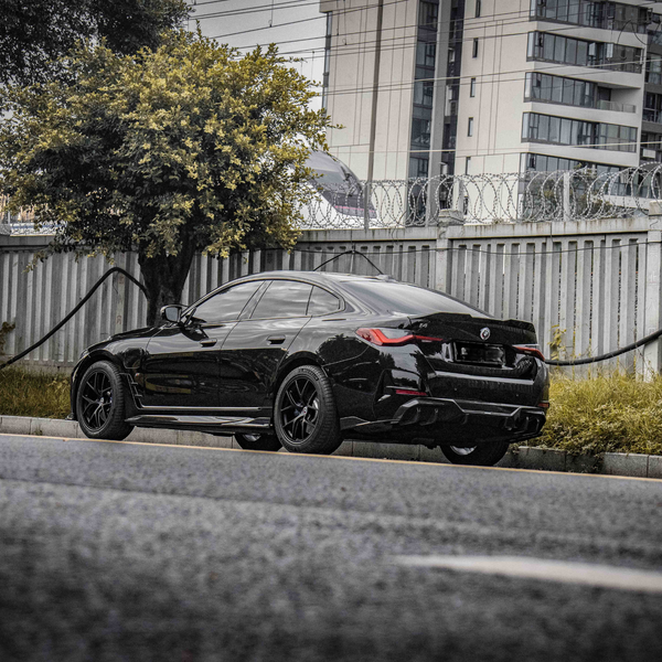 A sleek black car is parked alongside a street, with a tree and a metal fence topped with barbed wire in the background, and high-rise buildings beyond.