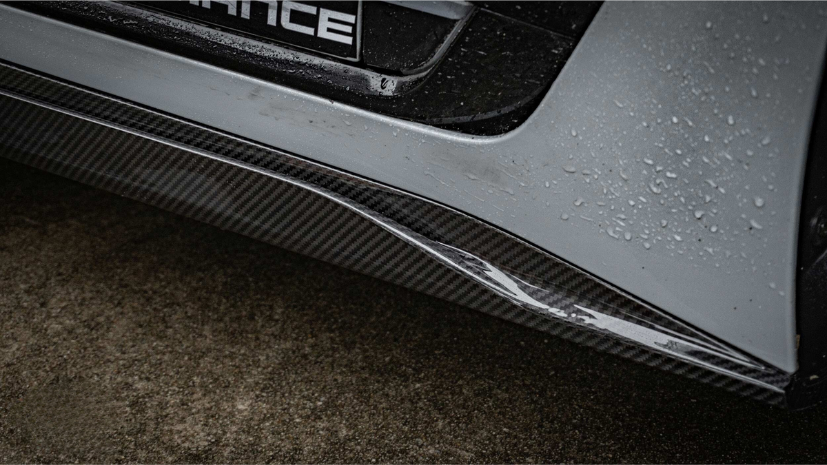 A carbon fiber car bumper with rain droplets glistening is shown against a wet pavement. Part of the word "ANCE" is visible on a nearby section.