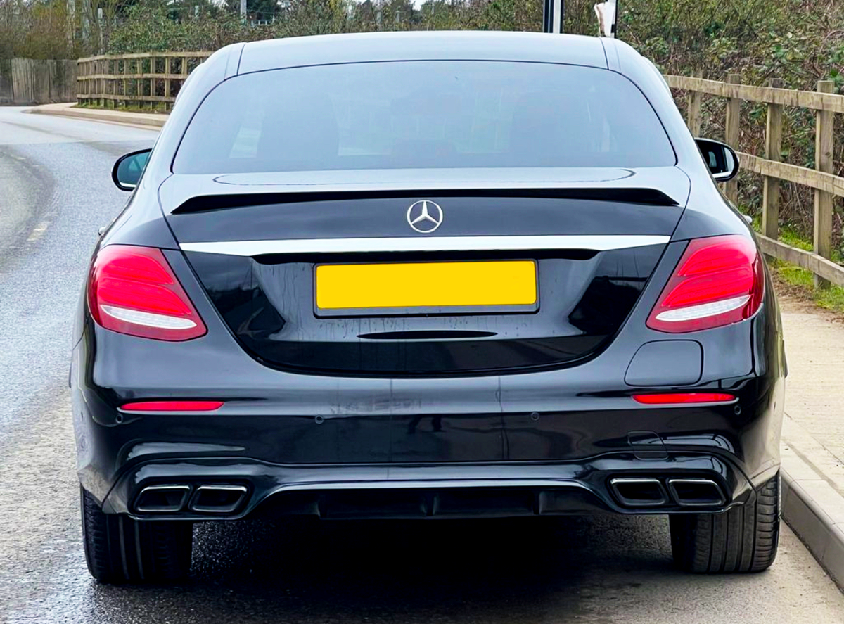 A black car is parked on a quiet road, aligned next to a wooden fence. The license plate is yellow, and a Mercedes-Benz logo is visible on the trunk.