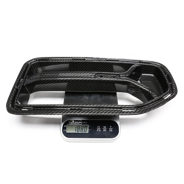 A carbon fiber car part rests on a digital scale, displaying "1180.0" grams. The scale has buttons labeled in another language, with a white background surrounding the setup.