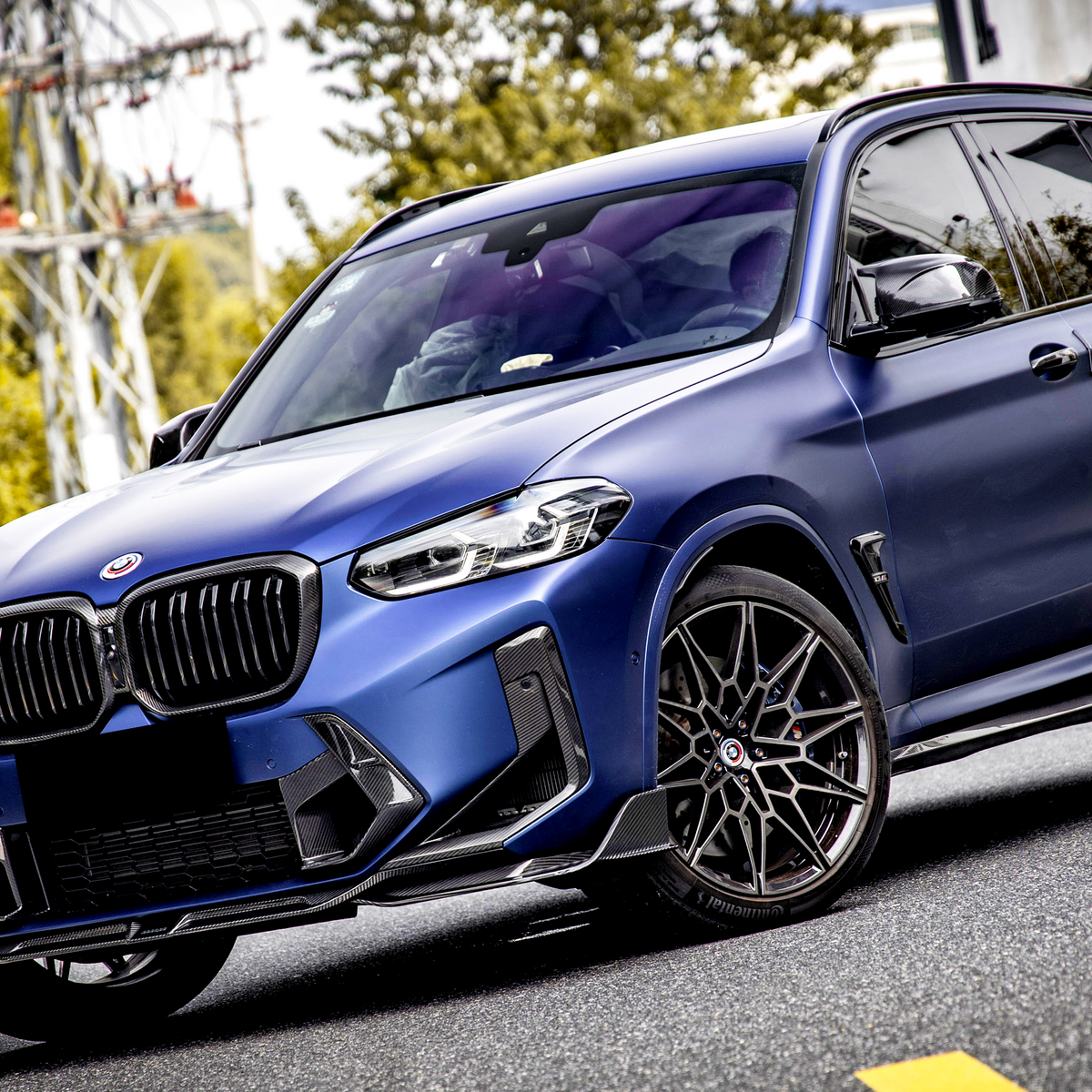 An SUV with a matte blue finish is parked on a street, featuring black grille and wheels, surrounded by trees and parked cars.