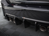 A car's black rear bumper with a diffuser features four exhaust pipes. It's parked on a patterned floor, reflecting light from above, creating a sleek and polished appearance.