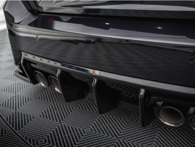 A car's black rear bumper with a diffuser features four exhaust pipes. It's parked on a patterned floor, reflecting light from above, creating a sleek and polished appearance.