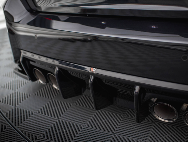 A car's black rear bumper with a diffuser features four exhaust pipes. It's parked on a patterned floor, reflecting light from above, creating a sleek and polished appearance.