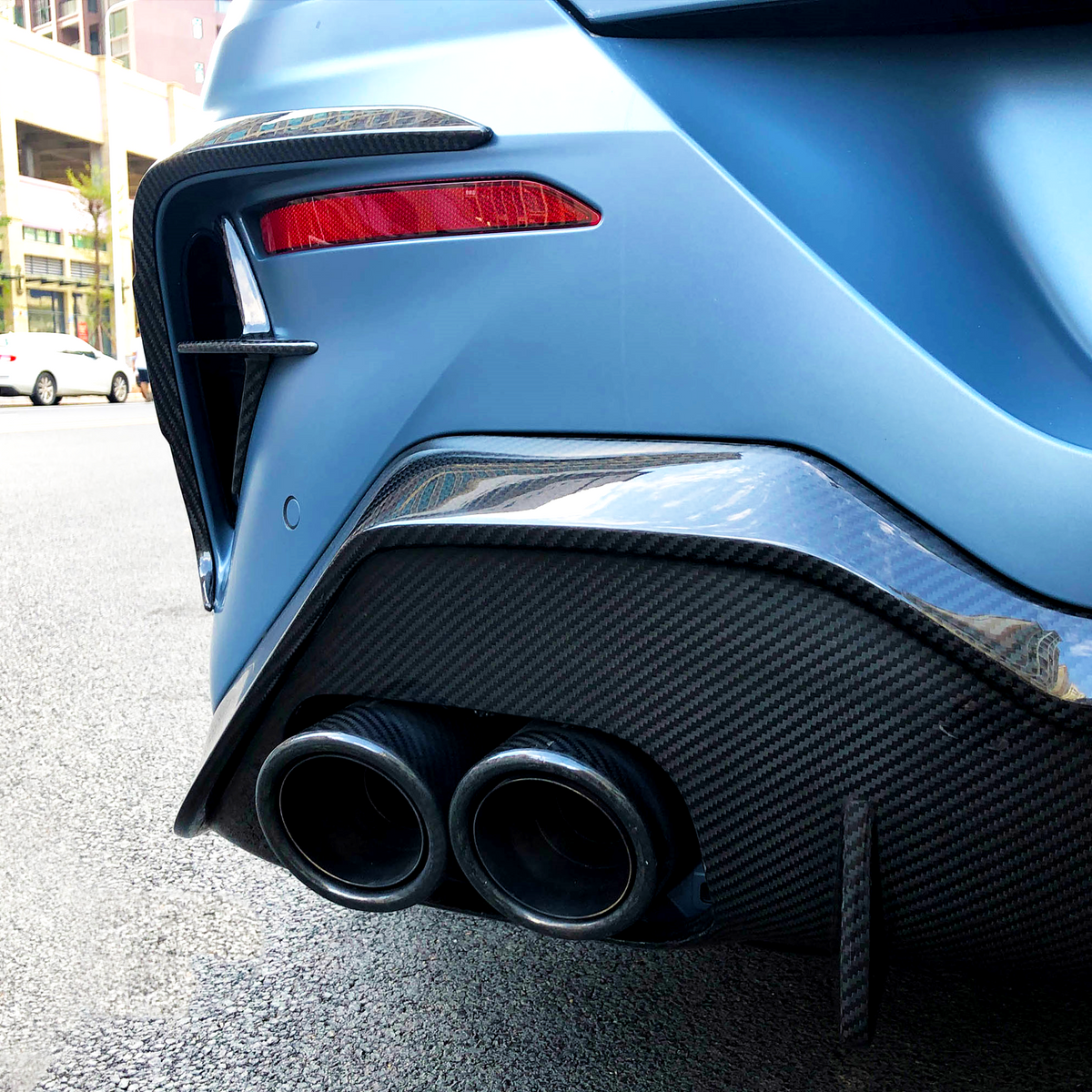 A blue car's dual exhaust pipes emit from a carbon fiber rear bumper on a city street, surrounded by parked white cars and a row of buildings.