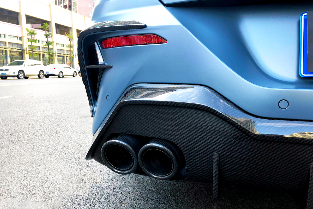 A blue car's dual exhaust pipes emit from a carbon fiber rear bumper on a city street, surrounded by parked white cars and a row of buildings.