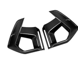 Two black carbon fiber car parts, shaped with angular edges, rest against a white background. They appear to be side mirrors or aerodynamic components.