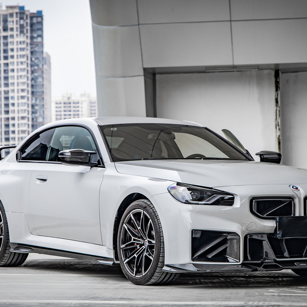 A sleek white car is parked in a rooftop garage, surrounded by tall city buildings. It has modern, angular bodywork and black detailing, emphasizing its sporty design.