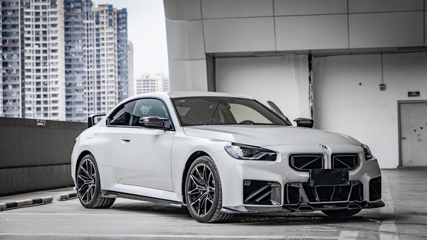 A sleek white car is parked in a rooftop garage, surrounded by tall city buildings. It has modern, angular bodywork and black detailing, emphasizing its sporty design.
