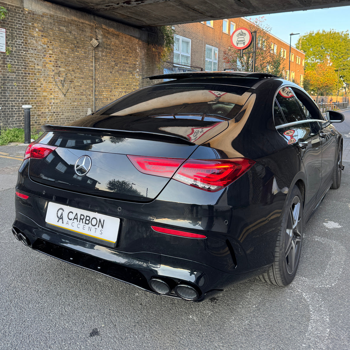 A black luxury car with a "Carbon Accents" plate is parked under a bridge. The surroundings include brick walls and buildings with trees under a clear sky.