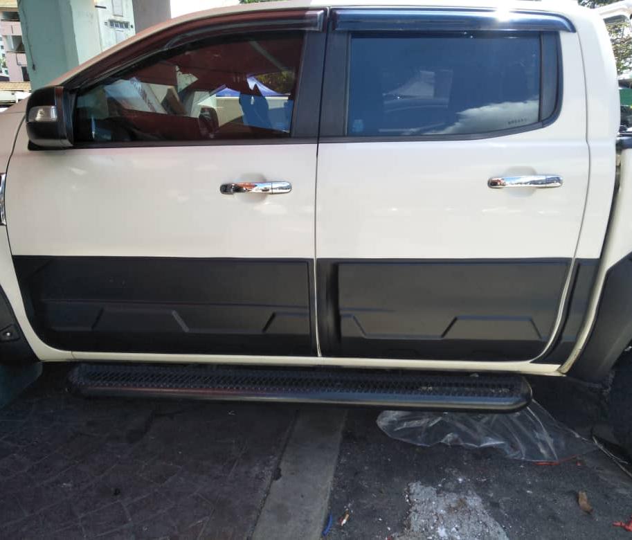 A white pickup truck is parked on a concrete surface, partially visible with its side doors and step board in focus. The background includes a building and other vehicles.