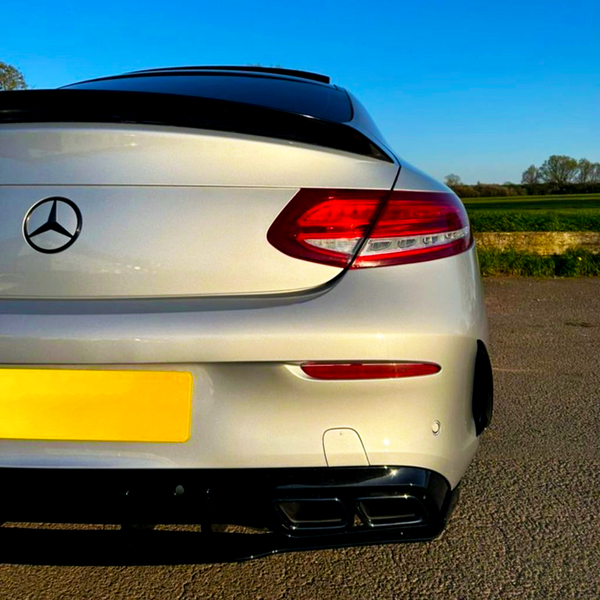 A white Mercedes-Benz car is parked on a paved surface, showcasing its rear end with red taillights under a clear blue sky, surrounded by green fields.