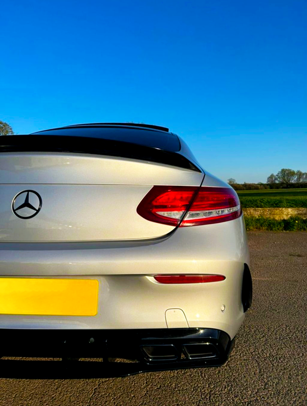 A white Mercedes-Benz car is parked on a paved surface, showcasing its rear end with red taillights under a clear blue sky, surrounded by green fields.