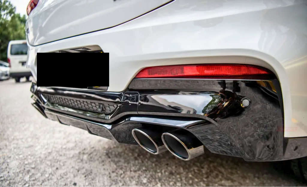 A white car's rear end is shown with dual exhaust pipes and a red taillight, parked on a gravel surface with another vehicle in the background.