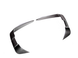 Two carbon fiber trim pieces, angled and sleek, lie on a plain white background, resembling modern automotive design elements, possibly for enhancing vehicle aerodynamics or styling.