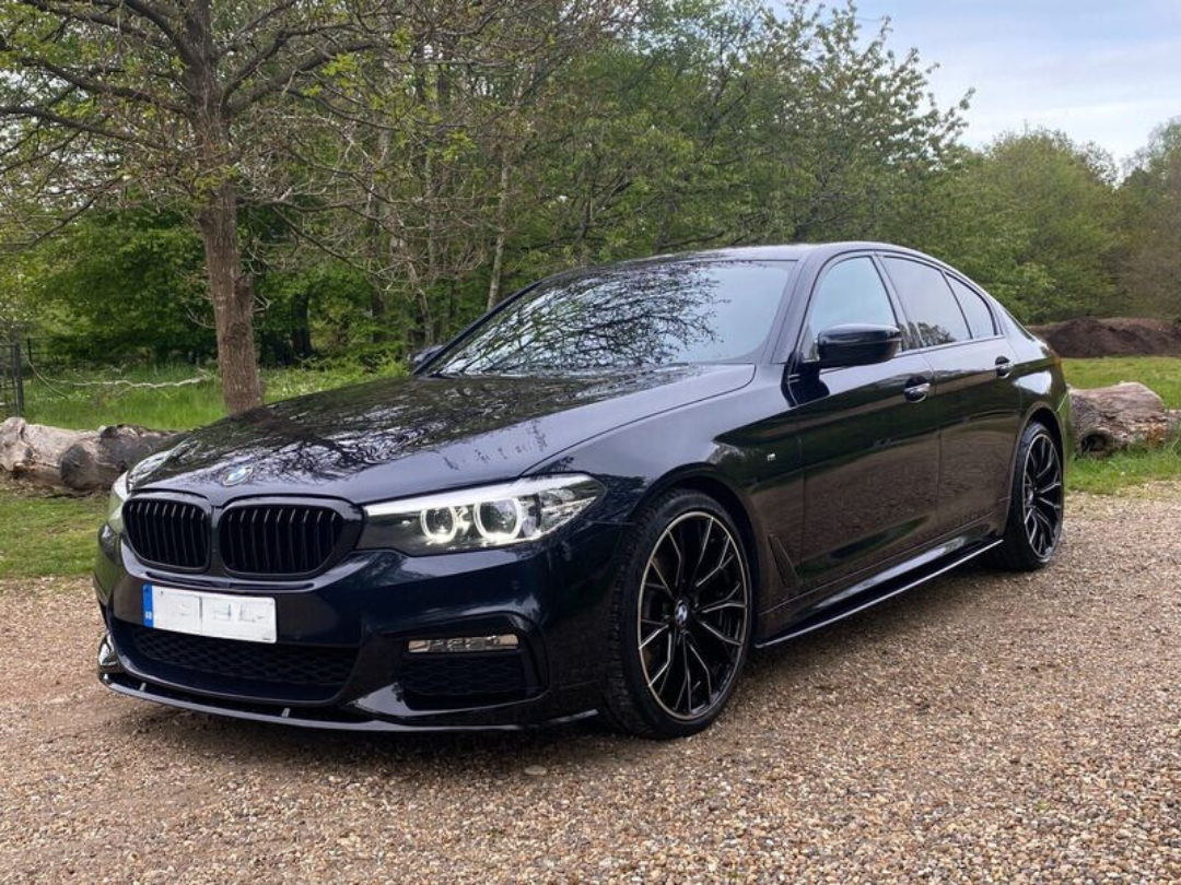 A black sedan with sleek design parked on a gravel path, surrounded by trees and greenery, under a clear sky. The car's headlights are on, reflecting the natural setting.