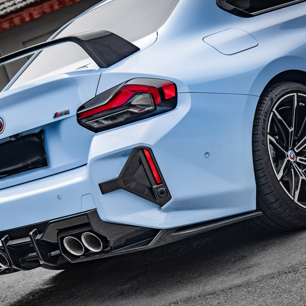 A light blue sports car displays a carbon fiber spoiler and quad exhausts, parked on an asphalt surface. The taillight design is angular and dramatic. Text on the bumper reads: "M2".