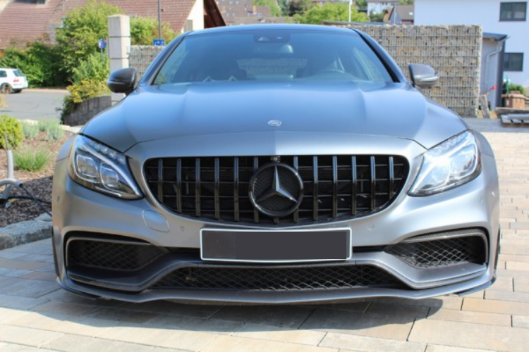 A silver car with a prominent grill, parked on a paved driveway. Residential houses and greenery are visible in the background on a sunny day.