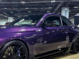 A purple sports car with sleek black wheels sits parked indoors at an automotive show, surrounded by a spacious industrial environment with overhead lighting and exposed ceiling structures.