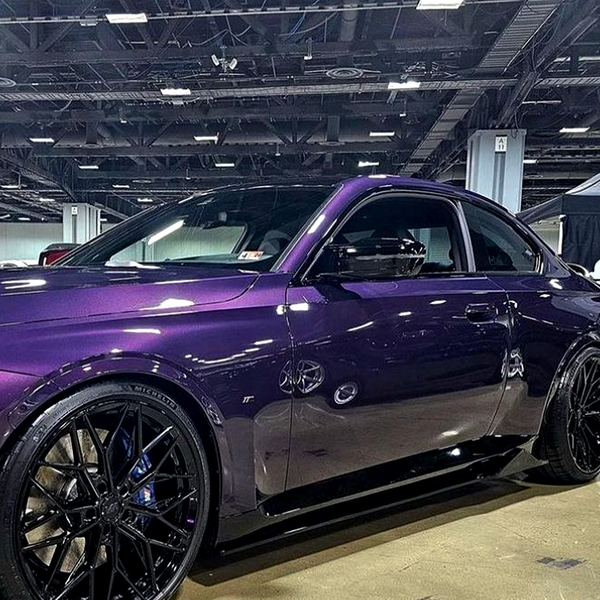 A purple sports car with sleek black wheels sits parked indoors at an automotive show, surrounded by a spacious industrial environment with overhead lighting and exposed ceiling structures.