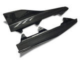 Two carbon fiber car side skirts resting on a white surface. The top piece features a stylized “TT” logo, enhancing its sleek, modern appearance.