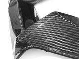 Carbon fiber car part with a glossy finish reflects light, highlighting its woven texture. It rests on a plain white surface, showcasing its sleek, angular design.