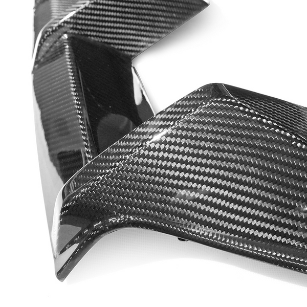 Carbon fiber car part with a glossy finish reflects light, highlighting its woven texture. It rests on a plain white surface, showcasing its sleek, angular design.