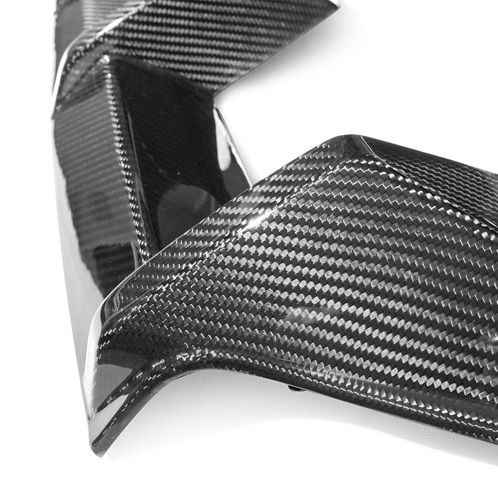 Carbon fiber car part with a glossy finish reflects light, highlighting its woven texture. It rests on a plain white surface, showcasing its sleek, angular design.