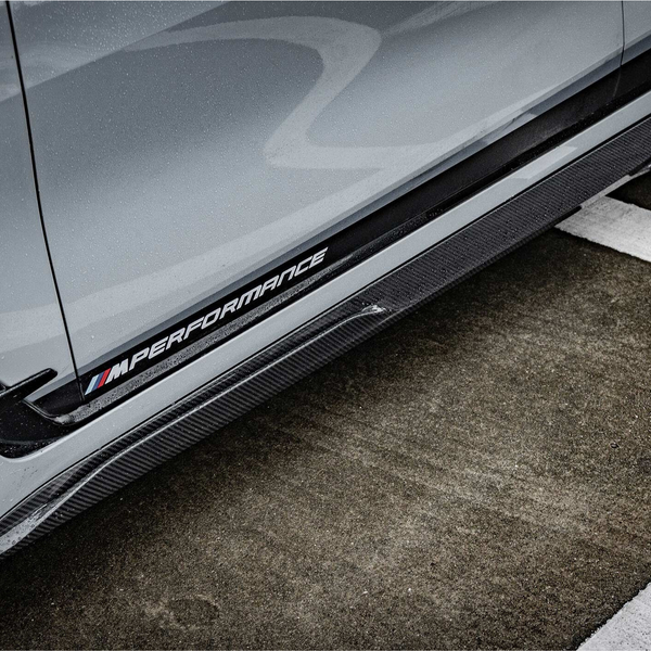 A gray car with an "M Performance" badge and aerodynamic features is parked on a wet, marked asphalt surface, highlighting its sporty design.