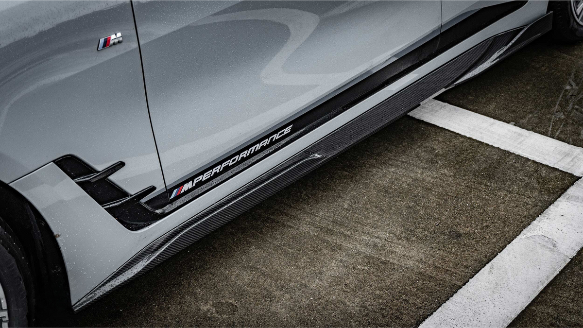 A gray car with an "M Performance" badge and aerodynamic features is parked on a wet, marked asphalt surface, highlighting its sporty design.