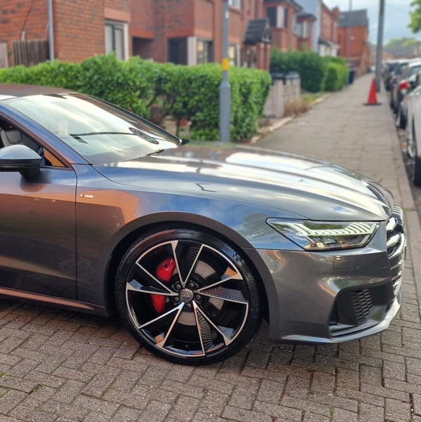 A sleek, silver car is parked on a brick driveway, showcasing its front wheel and headlight. Nearby, a residential street with red-brick buildings and greenery provides the setting.