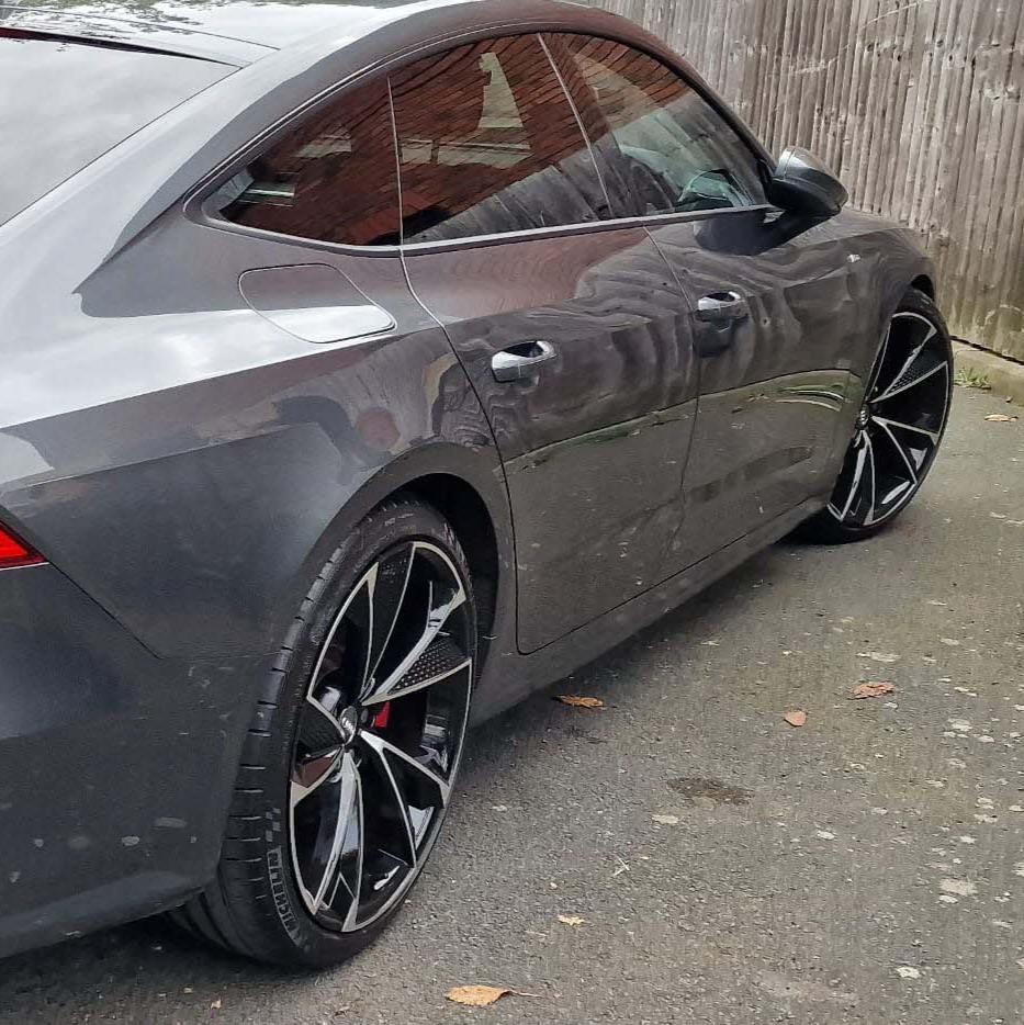 A sleek, gray car parked on a paved road, featuring shiny, black alloy wheels. It is adjacent to a wooden fence, with a few scattered leaves on the ground.