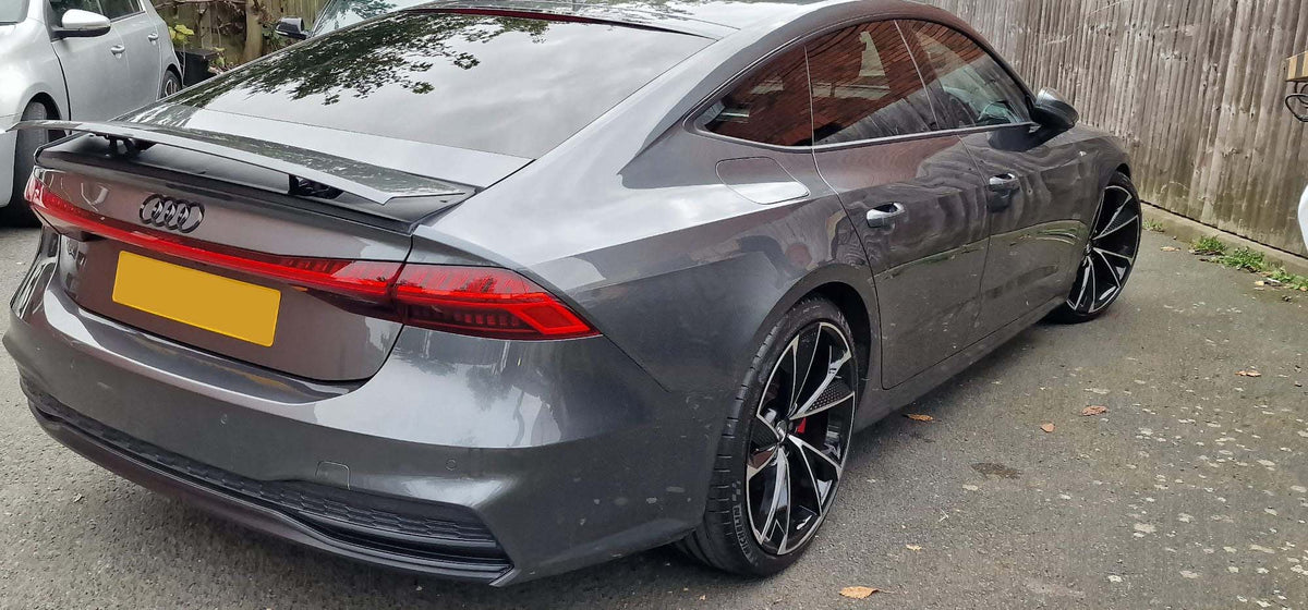 A gray Audi car is parked on a driveway beside a wooden fence. License plate is obscured, wheels are black with a unique design, and surroundings include another vehicle.
