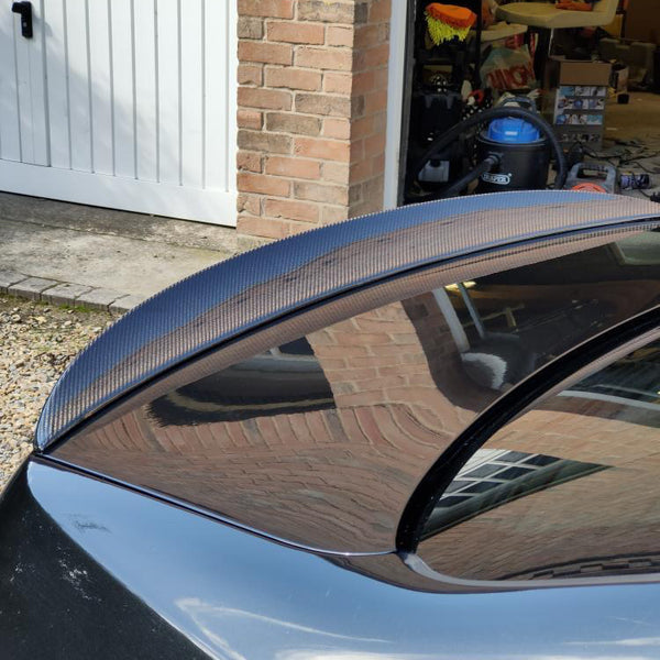 A black car with a carbon fiber spoiler is parked near a brick garage. The garage door is open, revealing various stored items, including a blue vacuum cleaner.