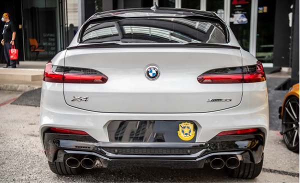 A white BMW X4 parked in front of a modern building with large glass doors. The car has dual exhausts and a badge labeled "xDrive 30i." There's a custom decorative plate.