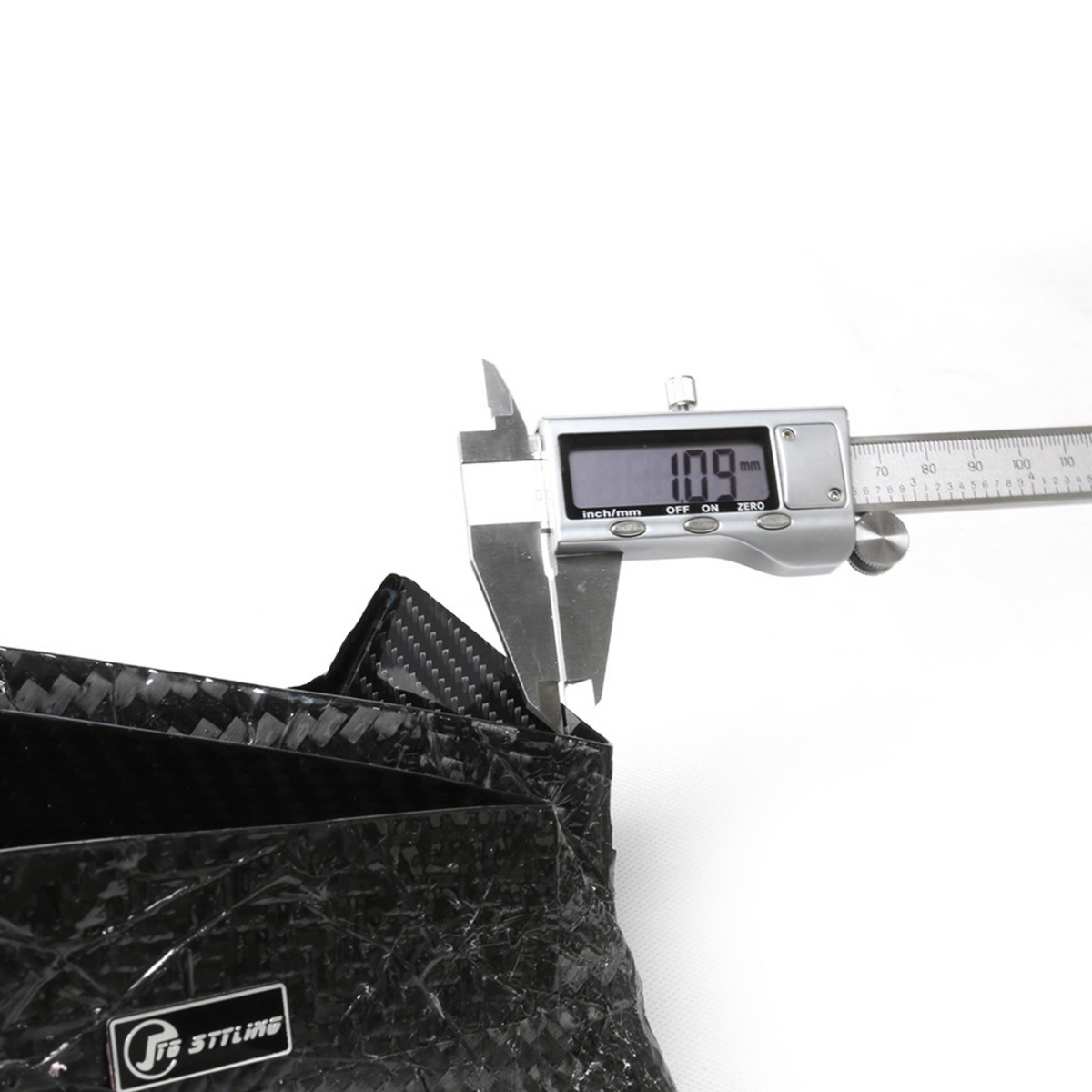 A digital caliper displays "10.9 mm" as it measures the thickness of a black carbon-fiber object. The setting is a clean, white background. Visible text: "Inch/mm OFF ON ZERO", "J's RACING".