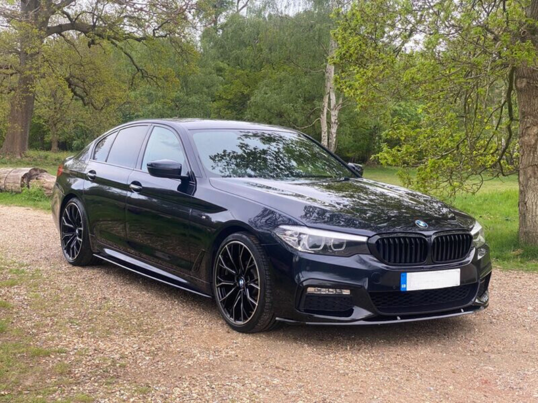 A black car is parked on a gravel path, surrounded by lush green trees and grass, creating a serene park-like setting.