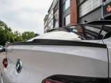 A white BMW car features a sleek rear spoiler, parked outdoors near a modern building with trees and more parked cars in the background.