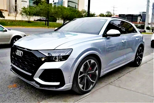 A silver SUV is parked on a city street. It features prominent branding on the front grille, sleek body lines, and large wheels. Nearby are trees and other vehicles.