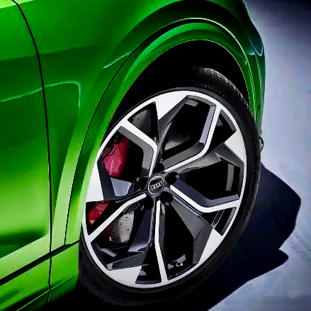 A shiny green car features a close-up of a stylish, geometric alloy wheel with a visible red brake caliper. The vehicle is positioned on a smooth, dark surface.