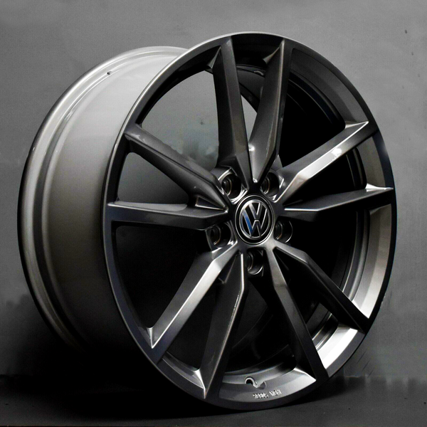 A dark metallic alloy wheel with a Volkswagen emblem rests on a plain surface. Bold, angular spokes create a sleek design. The setting is a simple gray backdrop.