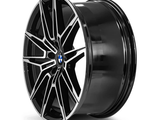 A black, multi-spoke alloy wheel with a central emblem sits in a plain, white background, highlighting its polished finish and intricate design.