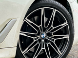 A car's wheel with a geometric alloy pattern and BMW logo is stationary on a paved surface, surrounded by part of the car's white fender and nearby ground.