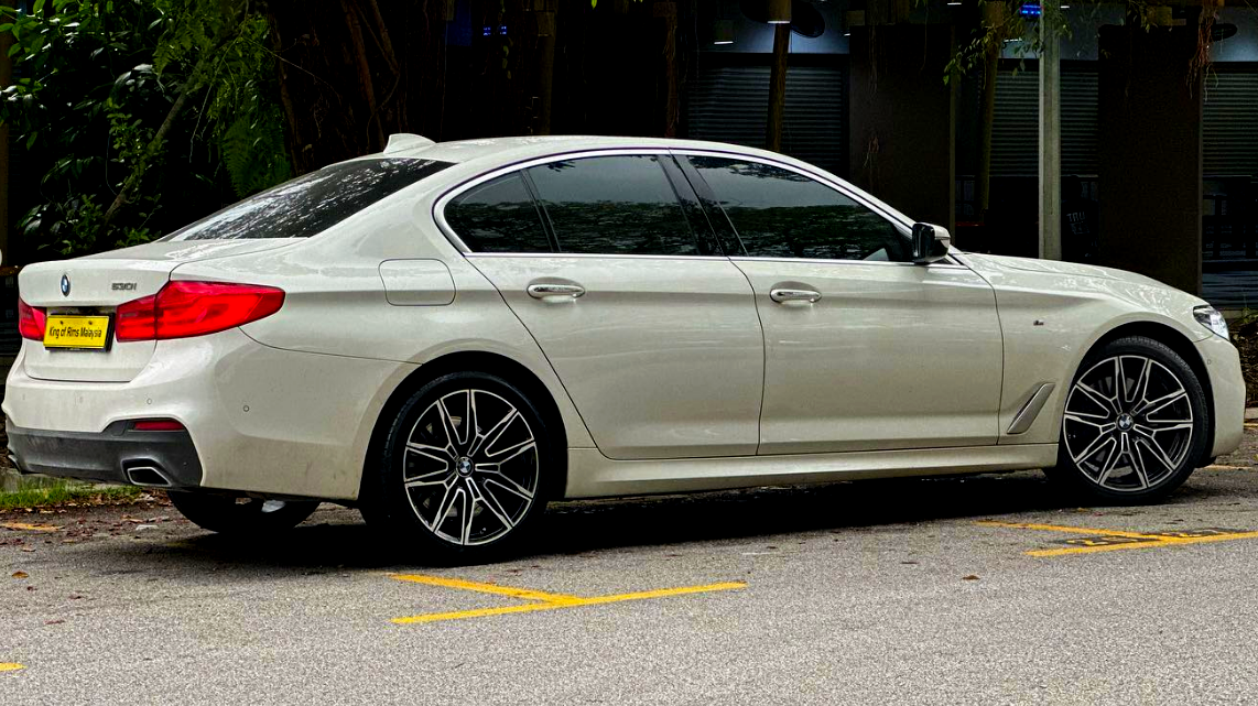A white BMW car is parked near trees, displaying sleek design and alloy wheels. The license plate reads "King of Rims Malaysia." Yellow parking lines are visible on the pavement.