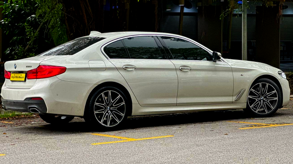 A white BMW car is parked near trees, displaying sleek design and alloy wheels. The license plate reads "King of Rims Malaysia." Yellow parking lines are visible on the pavement.