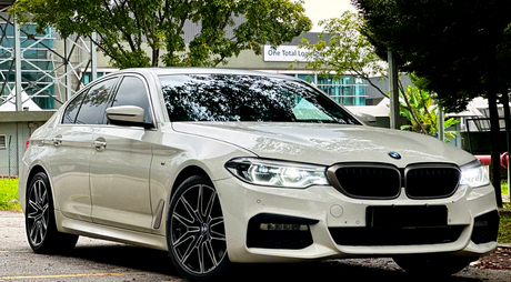 A white BMW sedan is parked on a street, surrounded by trees and near a modern building with a sign reading "One Total Logistics."