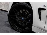 Black alloy wheel with BMW logo and M badge, stationary on a white car, parked on a patterned garage floor. The context shows part of the car's side panel and vent.