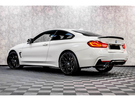A white BMW 430i coupe parked on a checkered floor with a textured wall background, showcasing sleek black rims and illuminated rear lights.